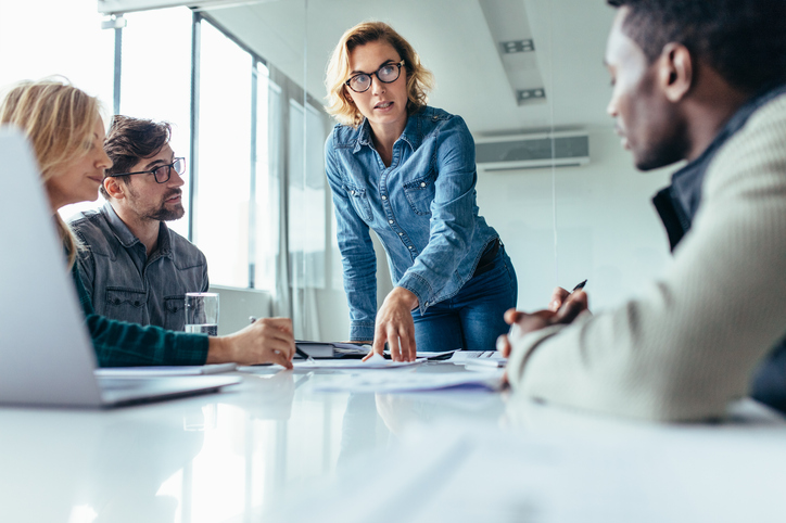 Business woman standing and speaking to coworkers who are sitting at a conference table.