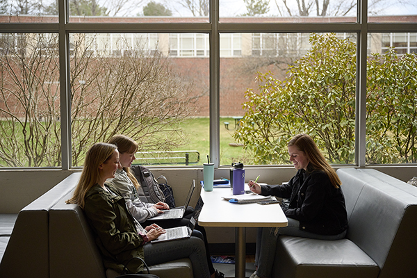 Three students sit at a table while writing on notepads and working on laptop computers.