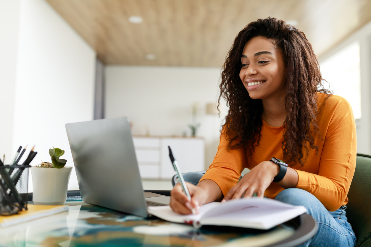 Smiling woman sits at a desk and works at a laptop while writing on paper.