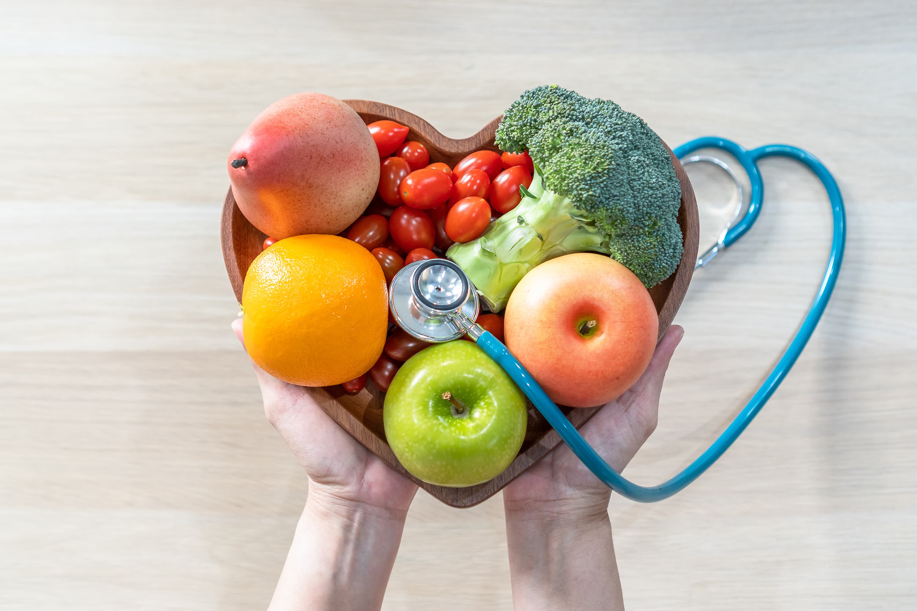 Hands holding a heart-shaped bowl with a stethoscope and fruit inside of it.