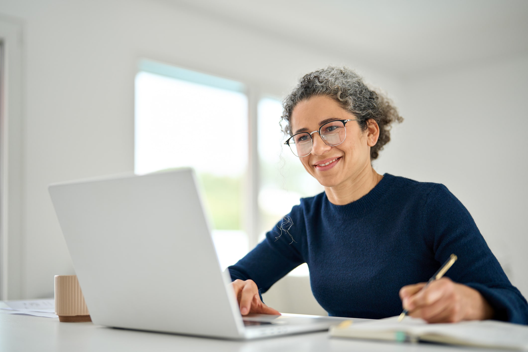 Smiling woman working on a laptop computer and writing in a journal.