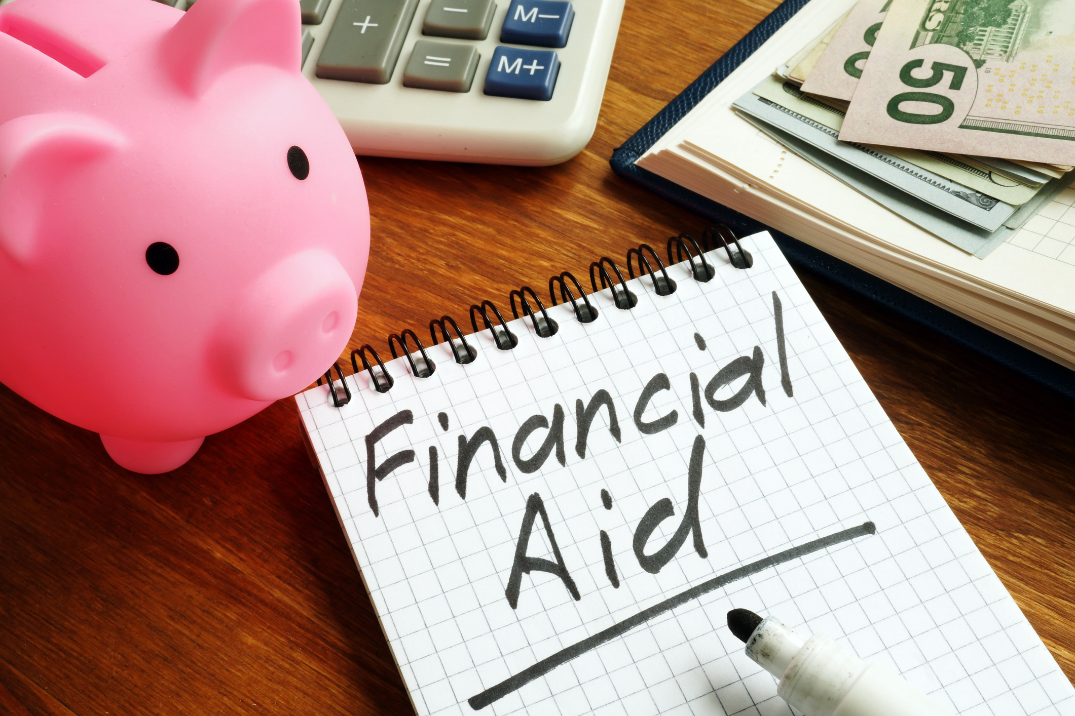 A financial aid sign, cash, piggy bank, and calculator sitting on a wooden surface.